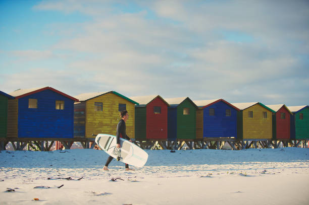 다채로운 해변 오두막이있는 서핑 보드와 함께 걷는 서퍼 - cape town beach hut multi colored 뉴스 사진 이미지