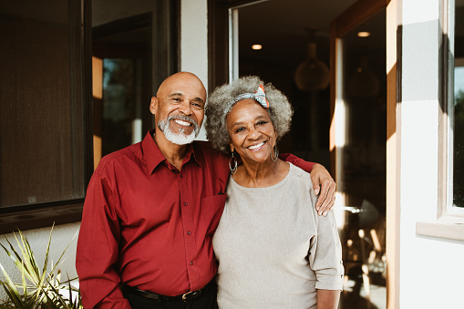 Portrait of retired man standing with arm around wife at front yard. Smiling senior couple is against house. They are in casuals.