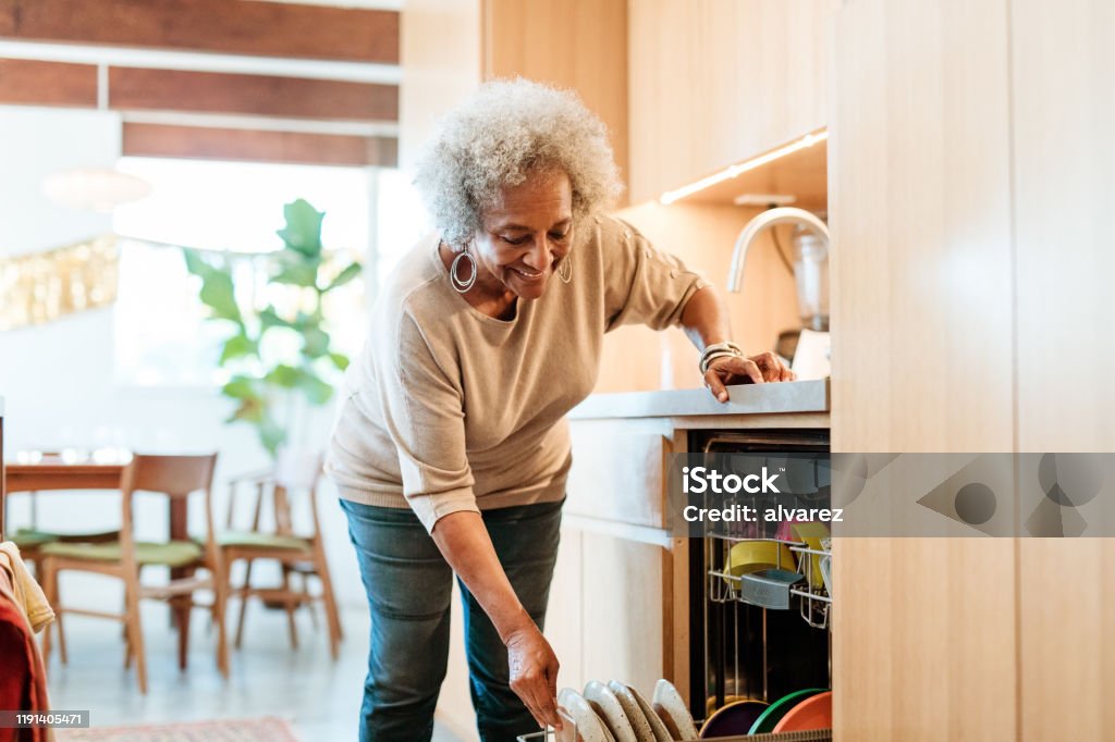 Smiling senior woman keeping plates in dishwasher Smiling senior woman keeping plates in dishwasher. Retired elderly female is doing routine chores in kitchen. She is in casuals at home. Senior Adult Stock Photo