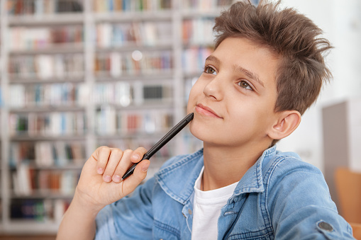 CLose up of a charming young boy enjoying studying at the library, looking away thoughtfully. Creativity, ideas, education concept