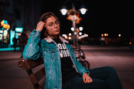 Portrait of a casual dressed teenage girl sitting on the bench in the city street and enjoying beautiful winter night