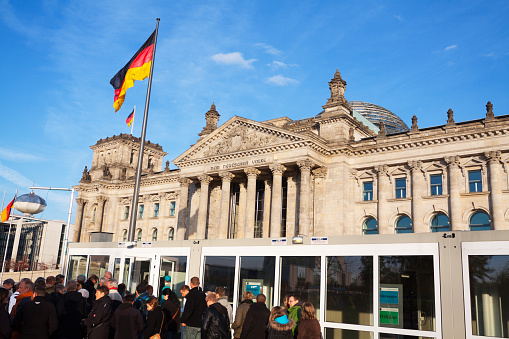 Queue of tourists at ticket counter for German Reichstag in Berlin with Reichstag in background. In foreground are many people and tourists applying for a ticket. Autumn season day. Between people is german flag in wind.