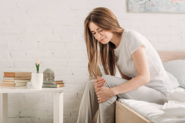 frowned girl sitting on bed near bedside table and suffering from knee pain stock photo