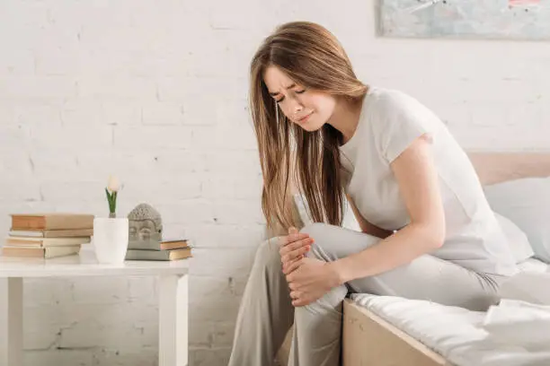 Photo of frowned girl sitting on bed near bedside table and suffering from knee pain