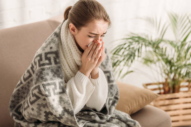 istock sick woman sneezing in napkin while sitting on sofa and wrapping in blanket 1191423761