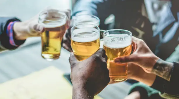 Photo of Group of multiracial friends enjoying a beer glasses in brewery english pub - Young people cheering at bar restaurant - Friendship and party concept - Focus on bottom black hand