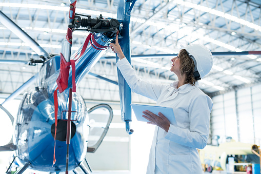 An engineer woman appears in the image inside a ship with helicopters. He dressed in a white coat, appears at the back of a helicopter checking one of his propellers. In the hand he holds a tablet