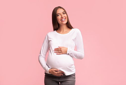 Pregnancy. Cheerful Pregnant Lady Touching Belly Smiling At Camera Standing On Pink Background. Studio Shot