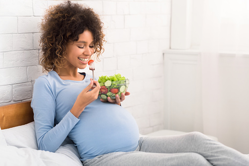 Happy pregnant woman eating natural vegetable salad, sitting on bed and having fresh snack, free space