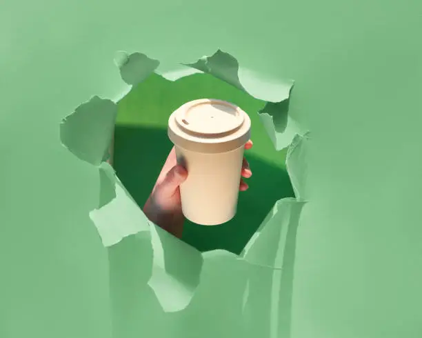 Hand holding a cup of coffee through a hole torn in neo mint color paper background. Get energy boost, hot drink in bamboo cup, travel eco friendly mug with lid. Copy-space on the cup.