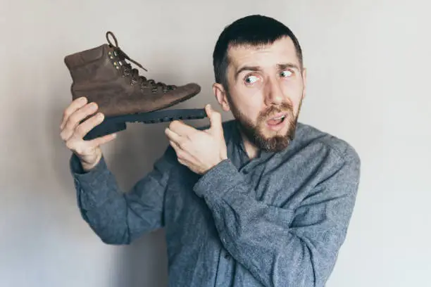 Photo of Man holding old leather boot with torn sole at his ear, surprise and shock on his face