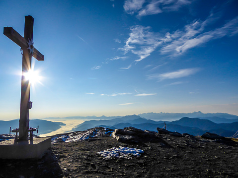 A metal cross on top of a mountain peak of Goldeck, Austria.  The cross is towering above another points. In the back there are some snow caped mountain chains. Serenity and calmness.