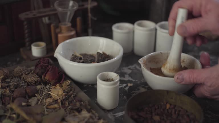 Close-up of table with dried flowers and vintage chemical containers. Old male Caucasian hands grinding powder using mortar and pestle. Ancient apothecary, alternative medicine, retro, healthcare.