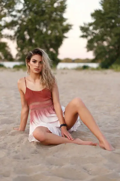 Portrait of a beautiful young girl on the beach. A girl in a light airy dress sits on the banks of the river. Model with large expressive eyes and puffy lips. Place for advertising.