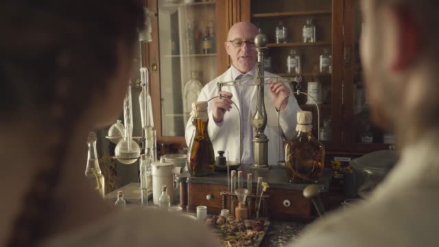 Mature Caucasian man standing at the table with vintage flasks, test tubes, vintage weighting machine, and talking. Senior guide working in retro pharmacy museum. Shooting over shoulder.