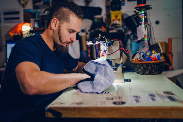istock Adult man tailor with beard sitting by his sewing machine at his work shop making a bag checking stitches handmade creative craft working at home 1191496366