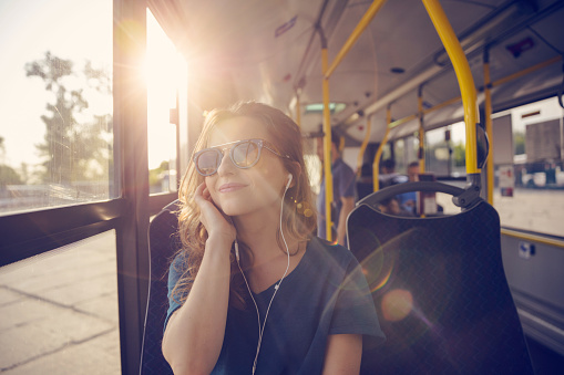 Smiling mid adult woman listening music in public transport. Happy female is commuting by bus on sunny day. She is wearing sunglasses.