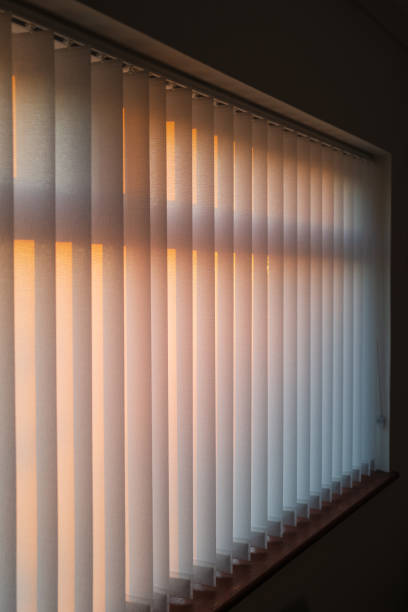 istock White vertical slat blinds hanging in front of a window as the sun is setting turning the light golden. The slats have sealed glued pockets and no cords at the bottom. Soft shallow focus 1191498521