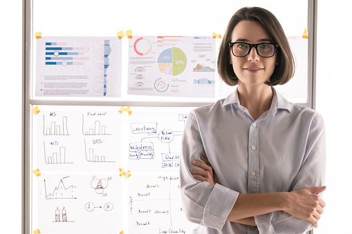 Pretty young brunette accountant or broker standing in front of camera against whiteboard with financial documents