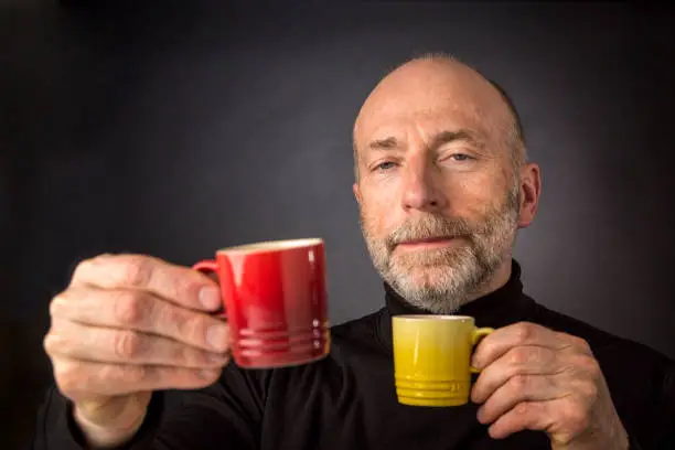 Morning coffee anybody? A senior bald man with a beard  is carrying two espresso coffee cups - a headshot against a black background