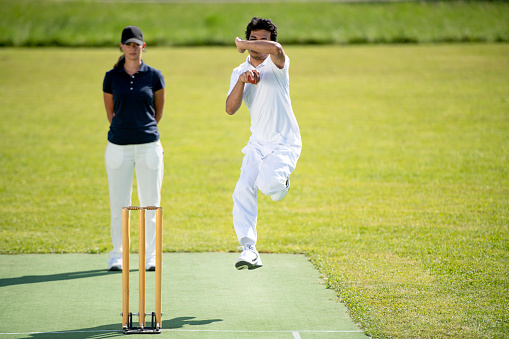 Photo of a cricket bowler in mid-air preparing to throw the ball. Umpire standing behind him.