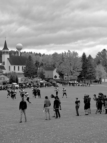 High school football game in “small town USA”, done in black and white. It is a homecoming game with the photo taken of Unrecognizable coaches and and high school football students from a distance, they are camera unaware and making a play while running down the field. The photo looks and feels like a rural small town scene complete with church and water tower in the background.