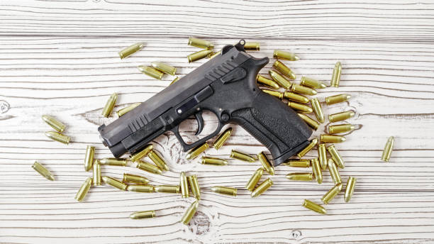 istock Top down view - modern black simple gun and yellow brass ammunition bullets scattered around on white wooden boards desk 1191540442