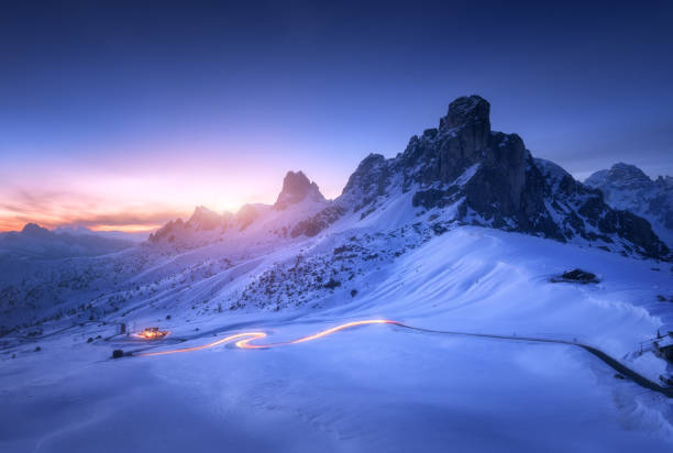 겨울에는 구불구불한 도로에서 눈 덮인 산과 흐릿한 자동차 헤드라이트. 눈으로 덮인 바위, 집, 산길, 푸른 별이 빛나는 하늘이있는 아름다운 풍경 돌로미티, 이탈리아 - cliff mountain winter snow 뉴스 사진 이미지