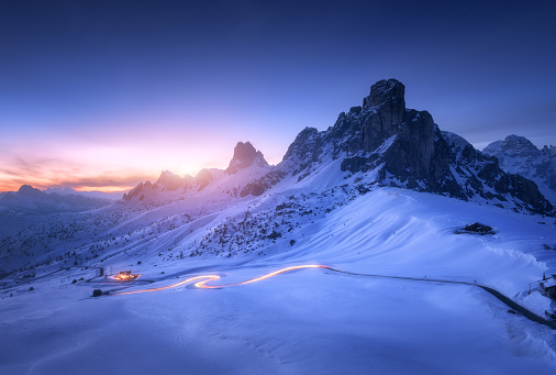 Snowy mountains and blurred car headlights on the winding road at night in winter. Beautiful landscape with snow covered rocks, house, mountain roadway, blue starry sky at sunset in Dolomites, Italy