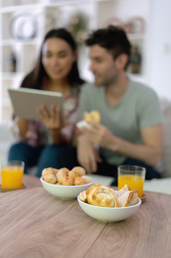 Focus on foreground of delicious empanadas and bread rolls while couple is looking at tablet sitting on couch - Lifestyles