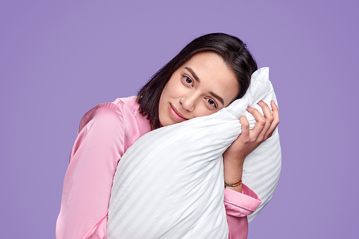 Pretty young female in pink nightwear hugging pillow while standing on purple background and looking at camera during bedtime