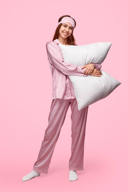 Cheerful young lady embracing pillow Full body happy young woman in silk pajama and sleep mask smiling and hugging soft pillow while standing against pink background during bedtime pyjamas stock pictures, royalty-free photos & images