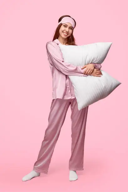 Full body happy young woman in silk pajama and sleep mask smiling and hugging soft pillow while standing against pink background during bedtime