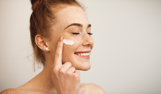 Close up of a young female with red hair and freckles applying white cream on her face laughing with closed eyes isolated on white background.