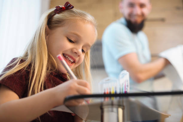 счастливая маленькая блондинка девушка улыбается во время рисования на кухне утром, пока ее отец мыть посуду . - family with one child domestic life caucasian love стоковые фото и изображения