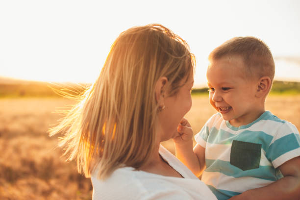 Close up portrait of lovely mother holding her little kid on arms and playing in a wheat field against sunset. stock photo