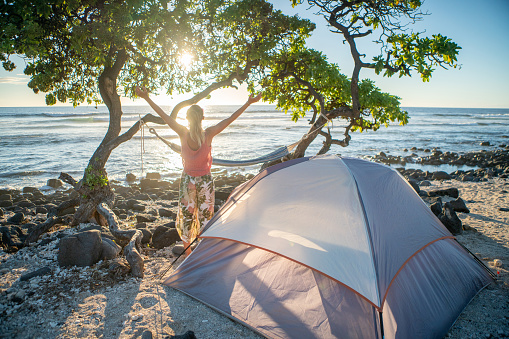 Girl camping on beach - Hawaii\nyoung woman camping on the beach stands arms wide open in front of tent at sunset
