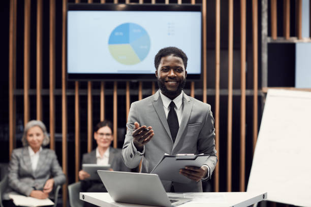 Portrait of smiling handsome young Afro-American manager with beard standing in conference room and using thesis while presenting budget information Smiling Afro-American manager presenting budget information dissertation stock pictures, royalty-free photos & images