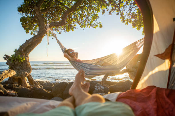 Point of view of man's feet from inside a tent camping on the beach in Hawaii looking at girlfriend in hammock outdoors stock photo
