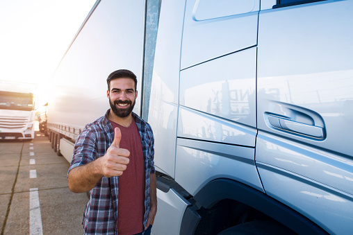 Portrait of young bearded man with thumbs up standing by his truck. Professional and positive truck driver standing by semi truck vehicle. Transportation services.