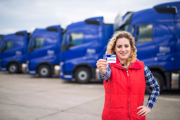 Commercial driving school. Truck driver showing driving license in front of truck vehicles. Woman truck driver proudly holding commercial driving license. In background parked trucks. Driving school and job openings for new drivers. driver's license stock pictures, royalty-free photos & images