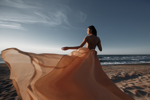 Girl in a light dress on the beach at sunrise.beautiful women in a light pink dress walking along the beach at dawn.Good morning and relaxation.Young beautiful girl standing on a sandy beach in a light pink dress on a background of blue ocean and sky