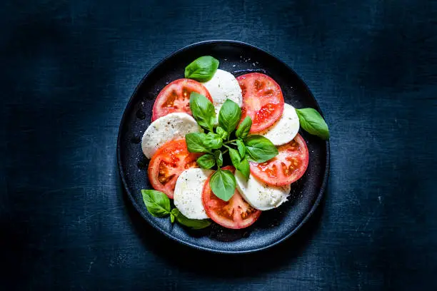 Top view of a caprese salad on a black plate. The salad is at the center of the image and is on a dark blackboard backdrop.  Low key DSLR photo taken with Canon EOS 6D Mark II and Canon EF 24-105 mm f/4L