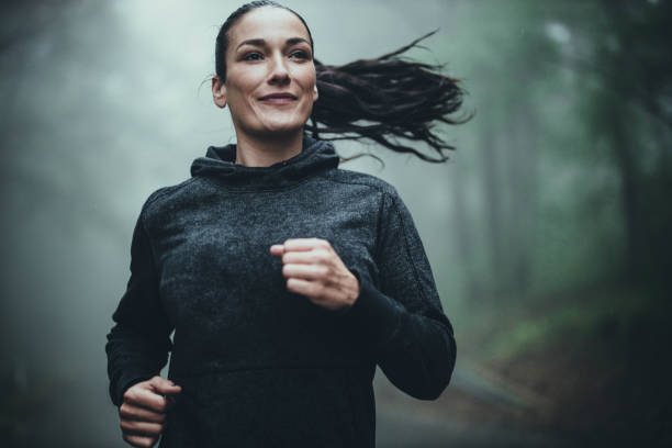 femme athlétique de sourire faisant pendant la journée brumeuse dans la forêt. - courir sous la pluie photos et images de collection