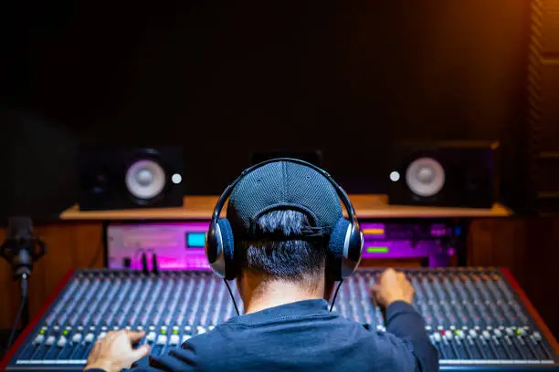 Photo of back of asian male professional music producer, sound engineer mixing a song on audio mixing console in recording studio. music production, post production concept