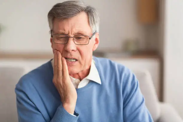 Elderly Man Having Toothache Touching Cheek Suffering From Pain Sitting On Sofa At Home. Selective Focus