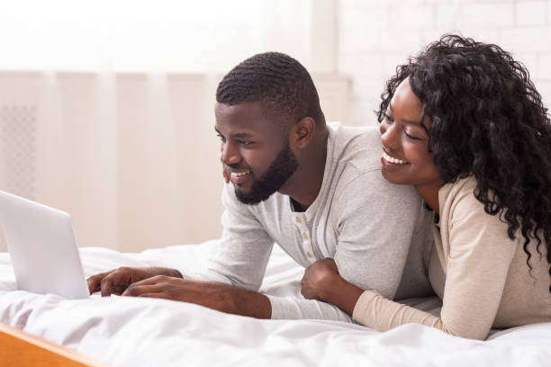 istock Cheerful afro spouses lying in bed using laptop together 1191647027