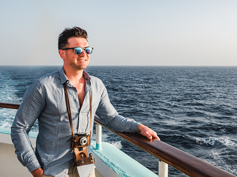 Fashionable man holding a vintage camera on the empty deck of a cruise liner against a background of sea waves. Side view, close-up. Concept of leisure and travel