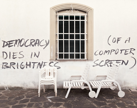 “Democracy dies in brightness (of a computer screen)“￼ is written on a wall, by a window with a metal grate. *** The text was digitally added and a release is provided ***