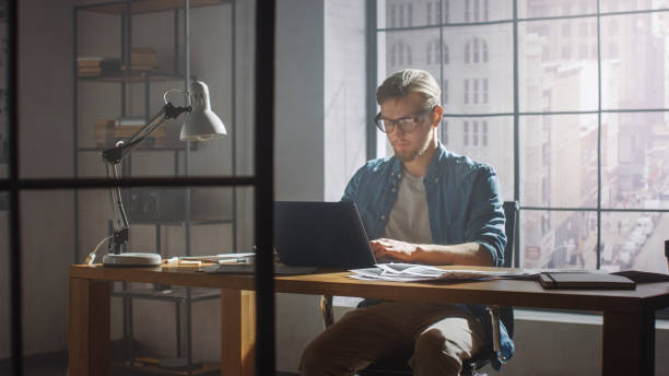 istock Creative Developer Works on a Laptop in His Studio. Designing Apps, Games and Doing Video Editing. In the Background Modern Sunny Loft Office with Project Storyboard and Concepts Covering the Wall 1191658308